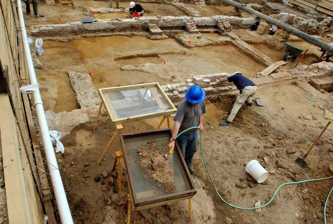 Archaeologists work among exposed brick and stone foundations, screening excavated soil through a mesh frame.